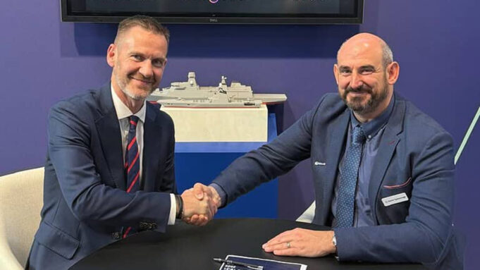 Babcock signs five-year engineering deal with AtkinsRealis Two men in blue suits shake hands across a table, smiling, with a model ship on a shelf behind them.