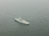 A gray military warship sailing in calm, open sea as seen from above.