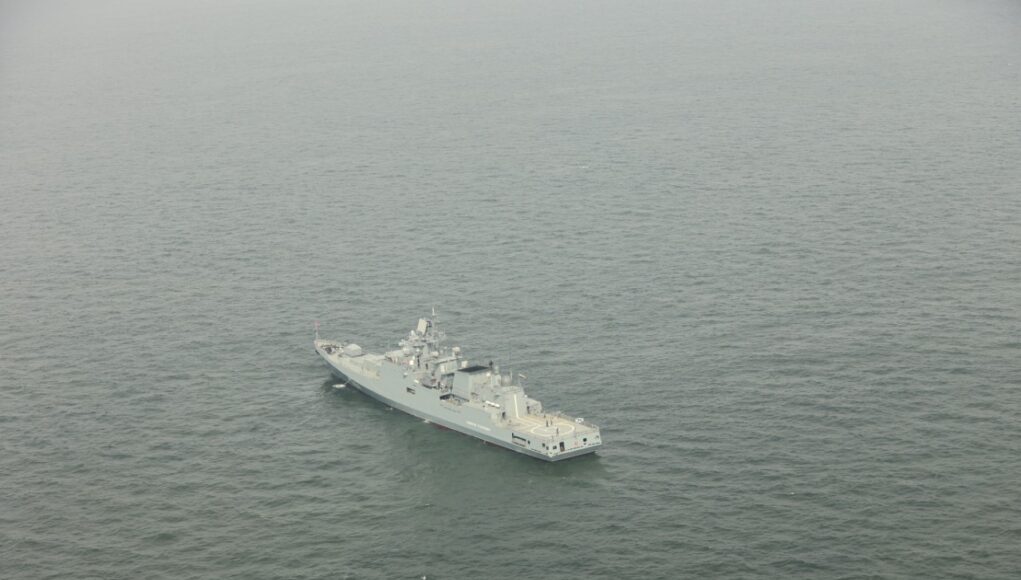 A gray military warship sailing in calm, open sea as seen from above.