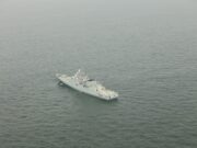 A gray military warship sailing in calm, open sea as seen from above.