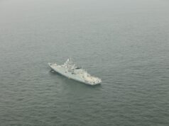 A gray military warship sailing in calm, open sea as seen from above.