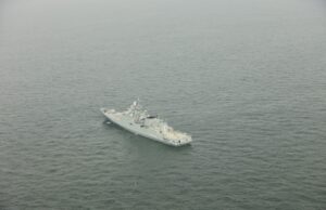 A gray military warship sailing in calm, open sea as seen from above.