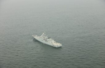 A gray military warship sailing in calm, open sea as seen from above.