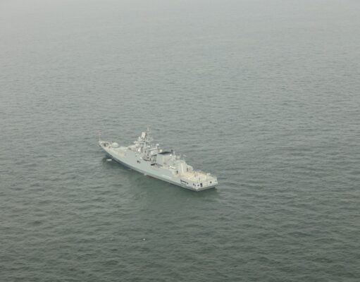 A gray military warship sailing in calm, open sea as seen from above.