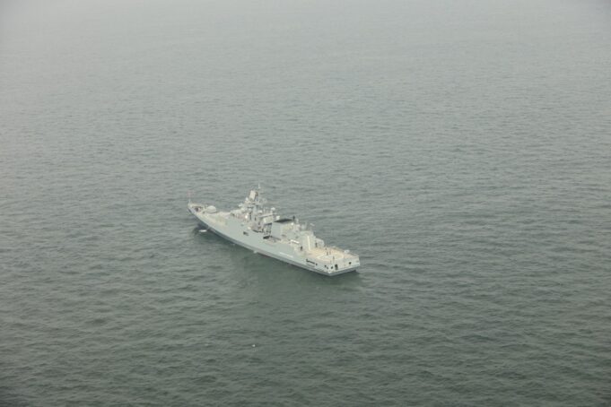 A gray military warship sailing in calm, open sea as seen from above.