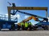 Team of workers in high-visibility jackets stand in front of blue scissor lift and yellow JCB boom lifts at an industrial yard, with a large yellow gantry crane labeled H & W in the background.