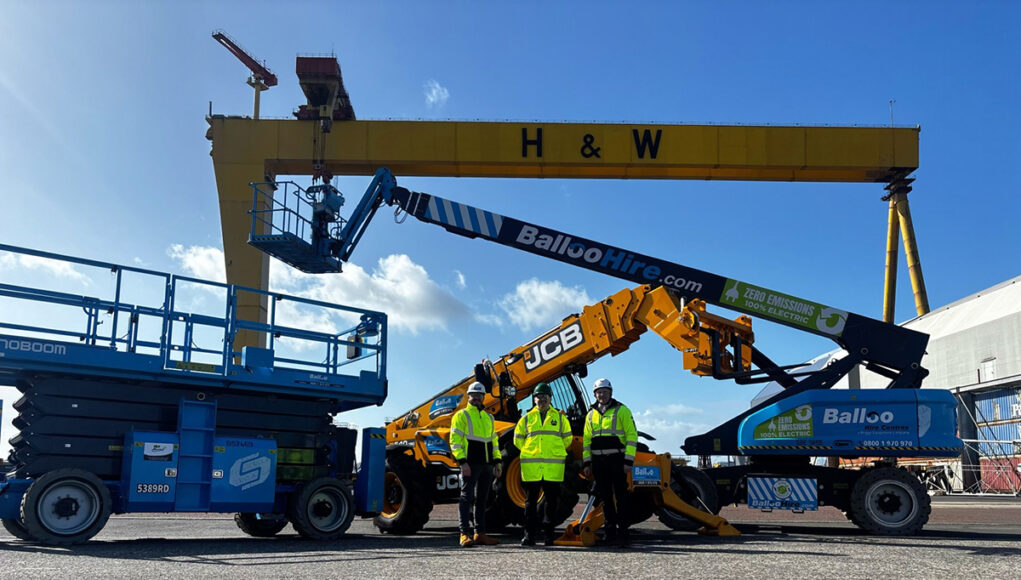 Team of workers in high-visibility jackets stand in front of blue scissor lift and yellow JCB boom lifts at an industrial yard, with a large yellow gantry crane labeled H & W in the background.