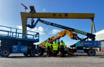 Team of workers in high-visibility jackets stand in front of blue scissor lift and yellow JCB boom lifts at an industrial yard, with a large yellow gantry crane labeled H & W in the background.
