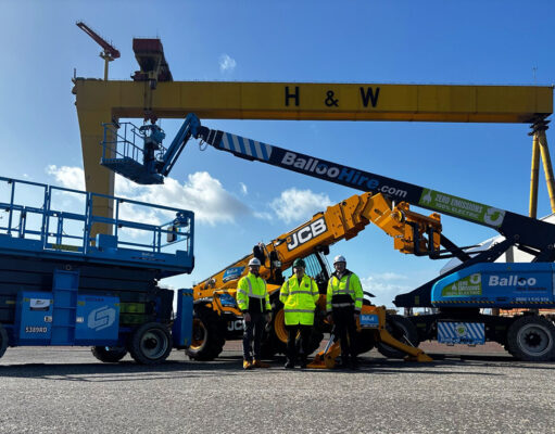 Team of workers in high-visibility jackets stand in front of blue scissor lift and yellow JCB boom lifts at an industrial yard, with a large yellow gantry crane labeled H & W in the background.