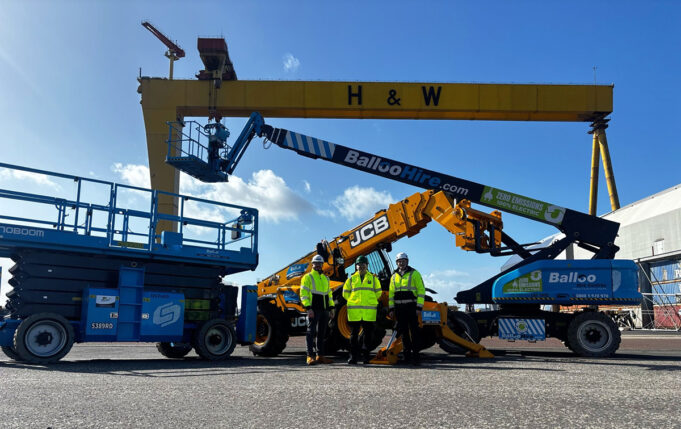 Team of workers in high-visibility jackets stand in front of blue scissor lift and yellow JCB boom lifts at an industrial yard, with a large yellow gantry crane labeled H & W in the background.