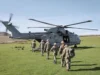 A Royal Navy helicopter on a grassy field with its side door open as soldiers in uniform walk toward it, carrying gear and backpacks.