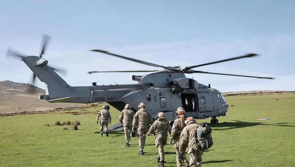 CHF-20260508-XA00314-010 A Royal Navy helicopter on a grassy field with its side door open as soldiers in uniform walk toward it, carrying gear and backpacks.