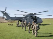 A Royal Navy helicopter on a grassy field with its side door open as soldiers in uniform walk toward it, carrying gear and backpacks.