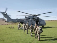 A Royal Navy helicopter on a grassy field with its side door open as soldiers in uniform walk toward it, carrying gear and backpacks.