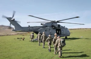 A Royal Navy helicopter on a grassy field with its side door open as soldiers in uniform walk toward it, carrying gear and backpacks.