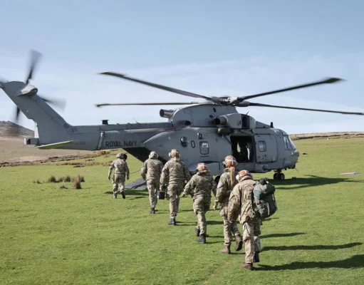 A Royal Navy helicopter on a grassy field with its side door open as soldiers in uniform walk toward it, carrying gear and backpacks.