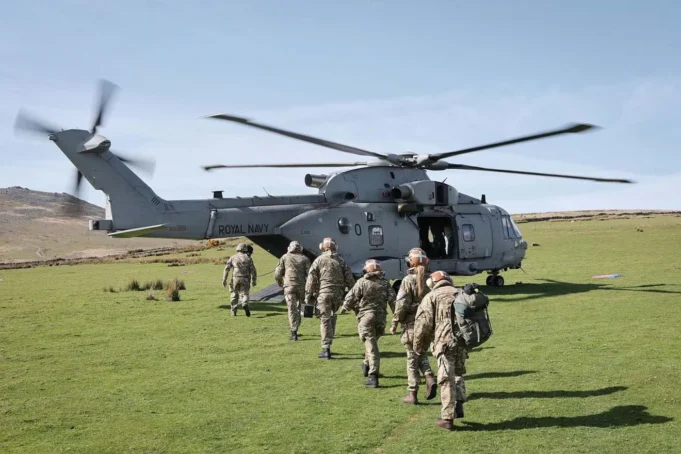 A Royal Navy helicopter on a grassy field with its side door open as soldiers in uniform walk toward it, carrying gear and backpacks.