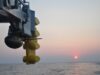 Close-up of a blue offshore platform component with yellow stabilizers hanging over a calm sea at sunset.