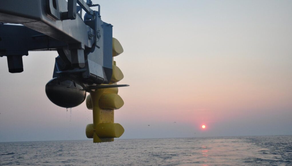 Close-up of a blue offshore platform component with yellow stabilizers hanging over a calm sea at sunset.
