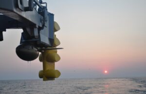 Close-up of a blue offshore platform component with yellow stabilizers hanging over a calm sea at sunset.