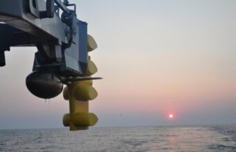 Canada picks Thales S2087 sonar for River-class ships Close-up of a blue offshore platform component with yellow stabilizers hanging over a calm sea at sunset.