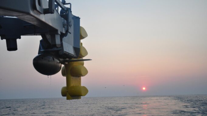 Canada picks Thales S2087 sonar for River-class ships Close-up of a blue offshore platform component with yellow stabilizers hanging over a calm sea at sunset.