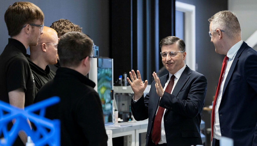 Group of professionals in a lab, a suited man explaining with hands raised to colleagues gathered around equipment.