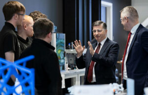 Group of professionals in a lab, a suited man explaining with hands raised to colleagues gathered around equipment.