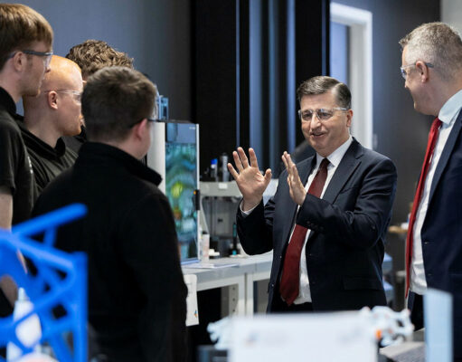 Alexander hails King’s Speech defence commitments Group of professionals in a lab, a suited man explaining with hands raised to colleagues gathered around equipment.