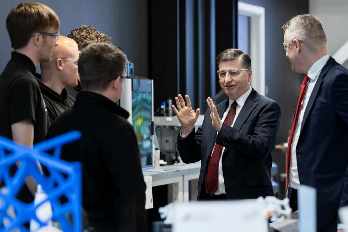 Alexander hails King’s Speech defence commitments Group of professionals in a lab, a suited man explaining with hands raised to colleagues gathered around equipment.