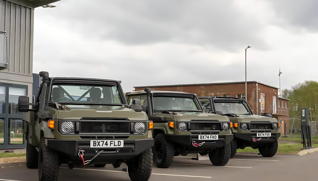 Row of olive green military-style Jeeps parked in a lot outside a building under a cloudy sky, front view shows license plates BX74 FLD and BX74 FEK.