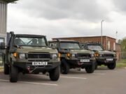 Row of olive green military-style Jeeps parked in a lot outside a building under a cloudy sky, front view shows license plates BX74 FLD and BX74 FEK.