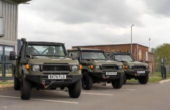 Row of olive green military-style Jeeps parked in a lot outside a building under a cloudy sky, front view shows license plates BX74 FLD and BX74 FEK.