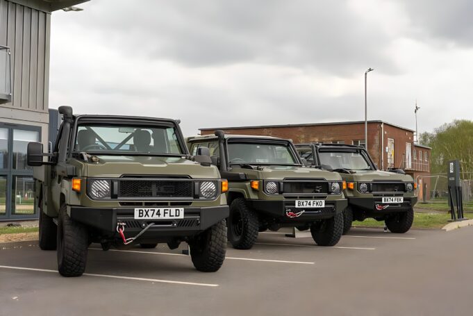 Row of olive green military-style Jeeps parked in a lot outside a building under a cloudy sky, front view shows license plates BX74 FLD and BX74 FEK.