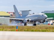 RAF transport aircraft lifting off from runway at an airbase with hangars and buildings in the background