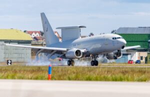 RAF transport aircraft lifting off from runway at an airbase with hangars and buildings in the background