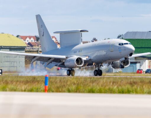 RAF transport aircraft lifting off from runway at an airbase with hangars and buildings in the background