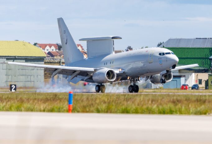 RAF transport aircraft lifting off from runway at an airbase with hangars and buildings in the background