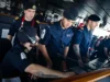 Royal Navy sailors in uniform operate a ship's bridge console, wearing headsets and focused on the controls.