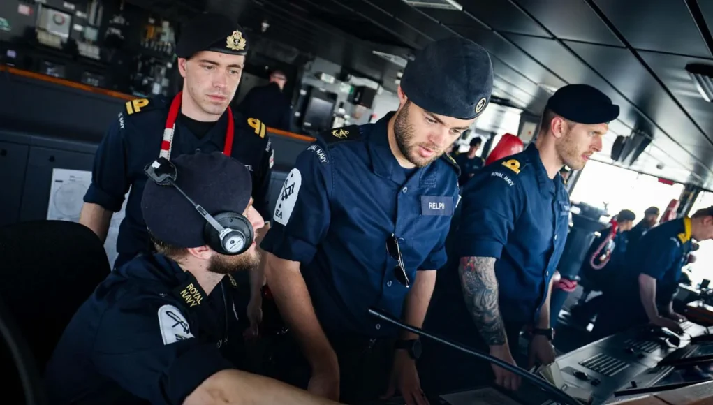 Royal Navy sailors in uniform operate a ship's bridge console, wearing headsets and focused on the controls.