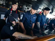 Royal Navy sailors in uniform operate a ship's bridge console, wearing headsets and focused on the controls.