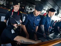 Royal Navy sailors in uniform operate a ship's bridge console, wearing headsets and focused on the controls.