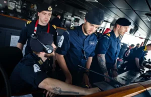 Royal Navy sailors in uniform operate a ship's bridge console, wearing headsets and focused on the controls.