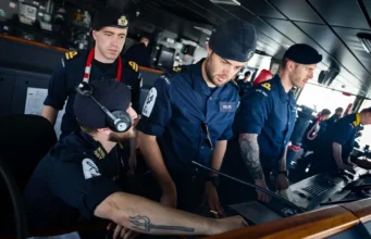 Royal Navy sailors in uniform operate a ship's bridge console, wearing headsets and focused on the controls.