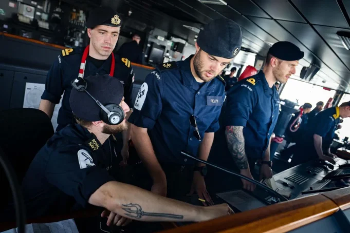 Royal Navy sailors in uniform operate a ship's bridge console, wearing headsets and focused on the controls.