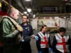 Team of workers in a shipyard or aircraft carrier hangar discussing work, with one officer in navy uniform and two women in reflective vests and gloves.