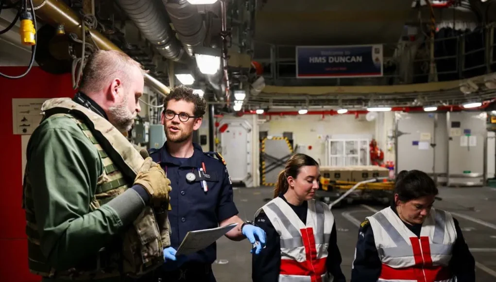 Team of workers in a shipyard or aircraft carrier hangar discussing work, with one officer in navy uniform and two women in reflective vests and gloves.