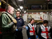 Team of workers in a shipyard or aircraft carrier hangar discussing work, with one officer in navy uniform and two women in reflective vests and gloves.
