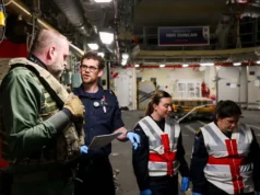 Team of workers in a shipyard or aircraft carrier hangar discussing work, with one officer in navy uniform and two women in reflective vests and gloves.