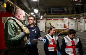 Team of workers in a shipyard or aircraft carrier hangar discussing work, with one officer in navy uniform and two women in reflective vests and gloves.